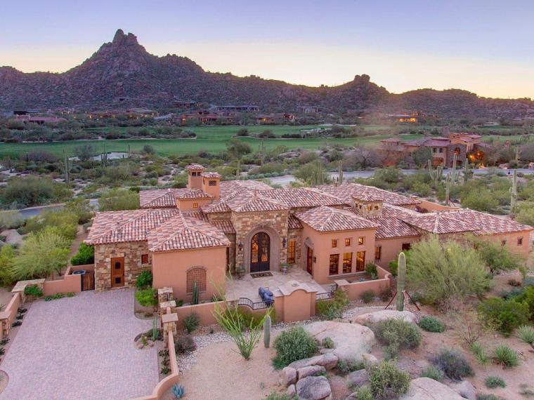 Aerial tile-roofed stone estate at dusk with a rocky peak and golf course behind