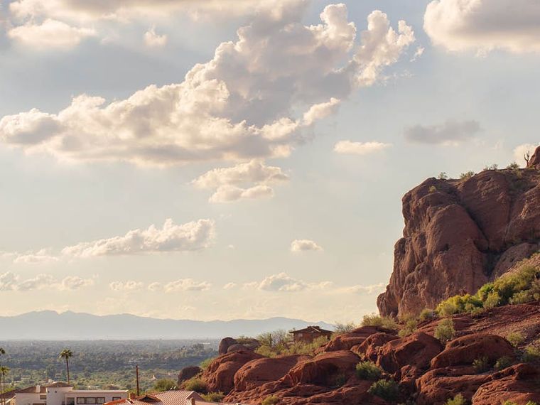 Red sandstone outcrop in the foreground above tile-roofed homes, the valley and distant ranges under tall bright cloud