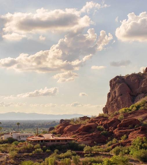 Red sandstone outcrop in the foreground above tile-roofed homes, the valley and distant ranges under tall bright cloud