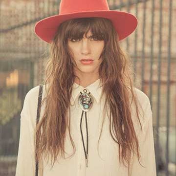 Woman in a wide-brim red hat and white blouse wearing a turquoise bolo tie, standing against a wrought-iron fence