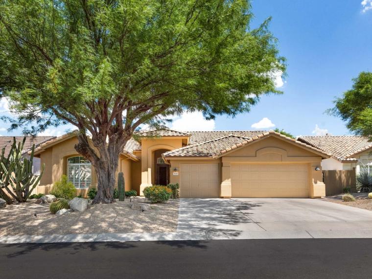 Single-story tan stucco house with tile roof and mesquite shading a gravel yard