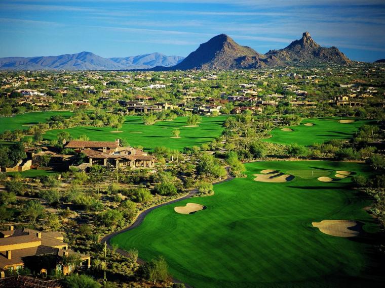 Elevated view over green fairways and bunkers threaded between tile-roofed homes, with a jagged mountain range on the horizon