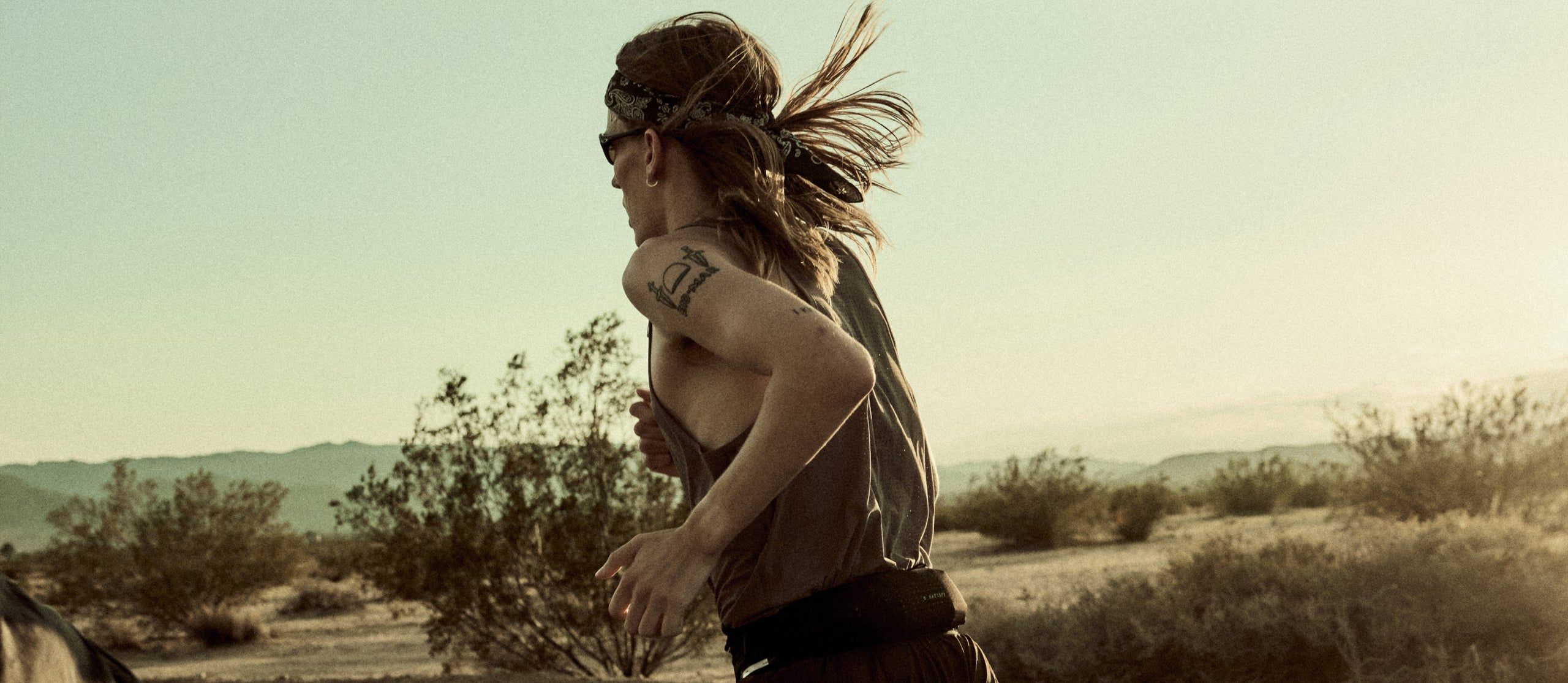 Satisfy Fall 26 — runner mid-stride on a desert highway at golden hour, in a Satisfy singlet and running shorts