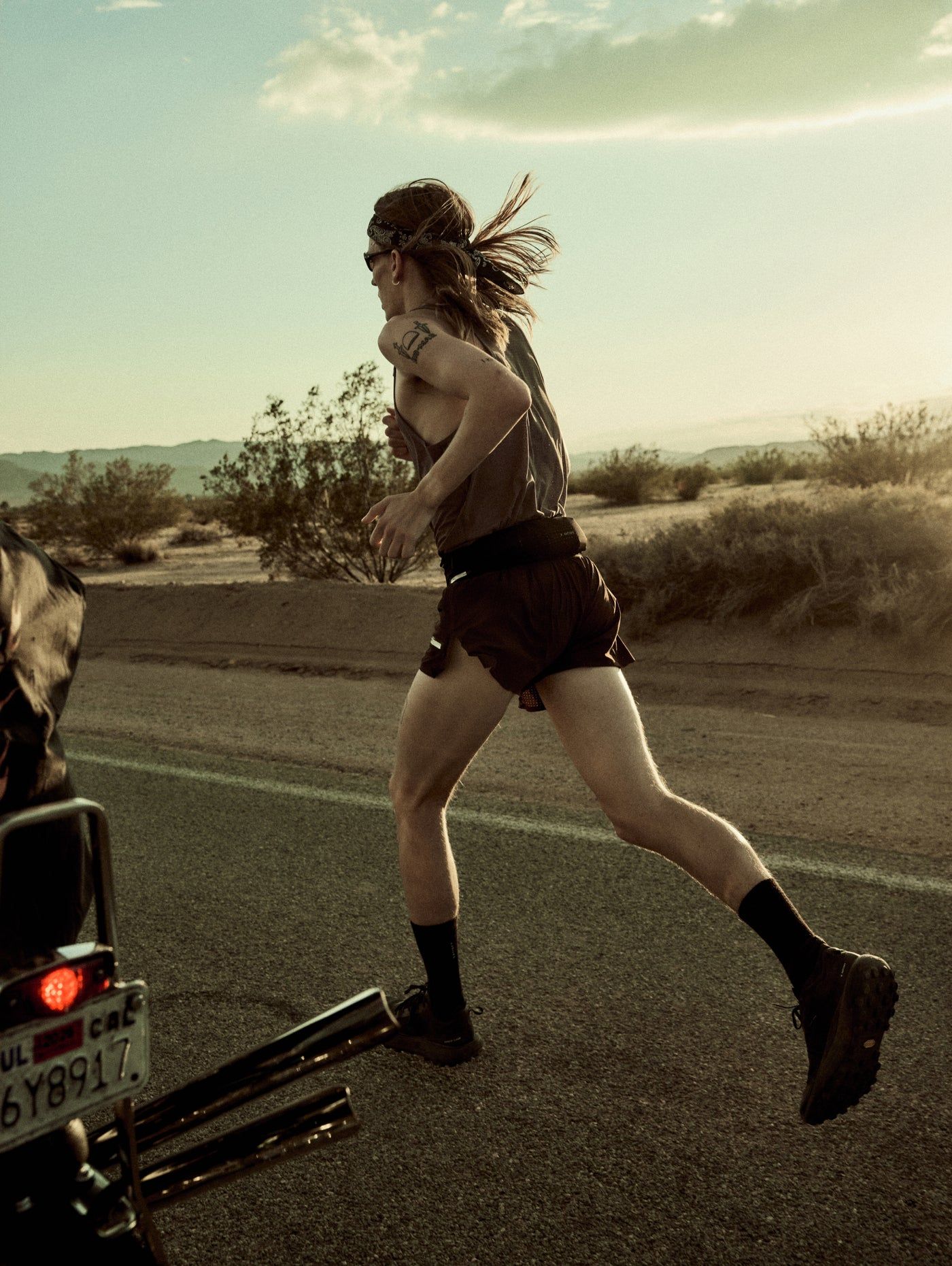 Satisfy Fall 26 — runner mid-stride on a desert highway at golden hour, in a Satisfy singlet and running shorts