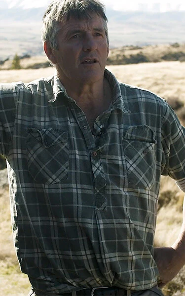 a man in a plaid shirt leans against a rock in a field