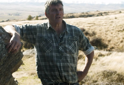 a man in a plaid shirt leans against a rock in a field