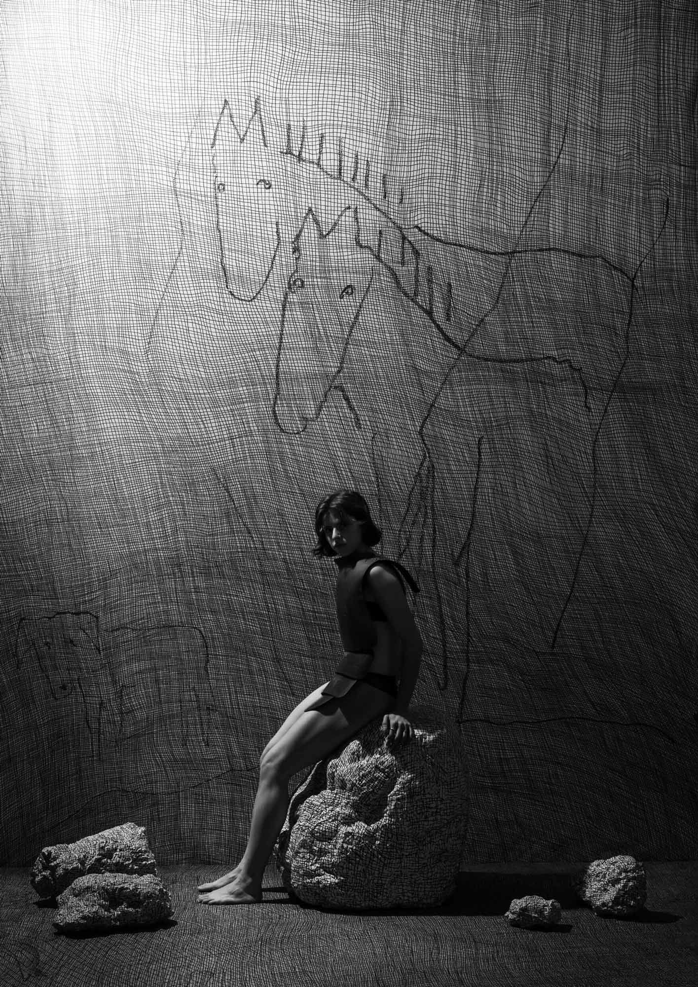 In a platinum print, a woman in armor-like attire sits on a boulder, illuminated from above, near an etched dual-horse mural.