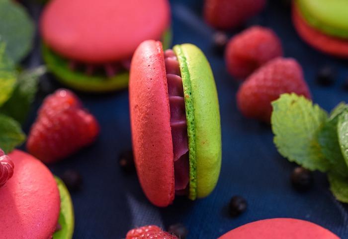 Bilde av mange makroner med rød topp og grønn bunn, med bringebær og mynteblad som ligger rundt.