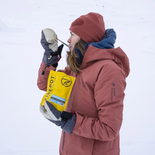En kvinne kledd i tykk vinterjakke, lue og votter spiser turmat rett fra posen