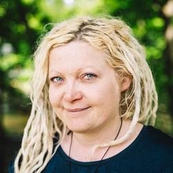 Smilende kvinne med platinablondt dreads-hår og blå øyne, med trær i bakgrunnen.