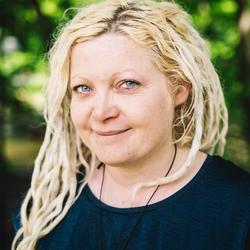 Smilende kvinne med platinablondt dreads-hår og blå øyne, med trær i bakgrunnen.