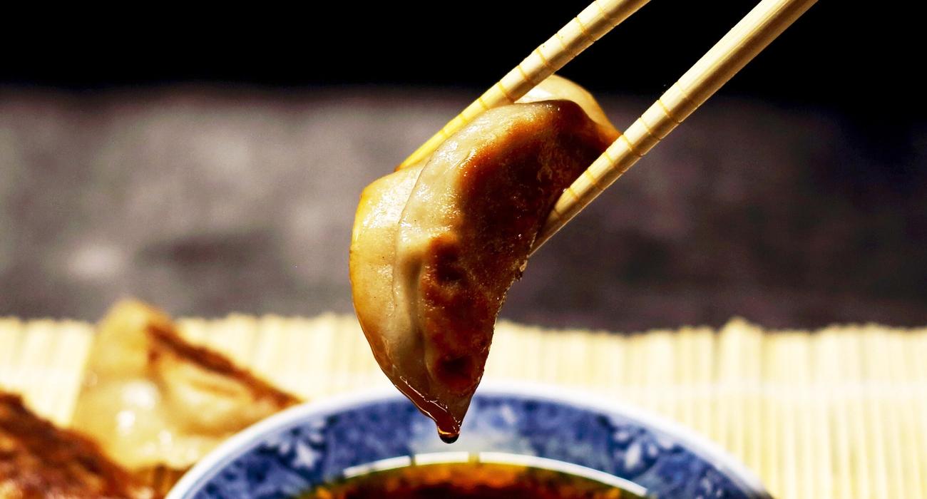 Bilde av en dumpling som blir holdt av to spisespinner og har blitt dyppet i soyasaus