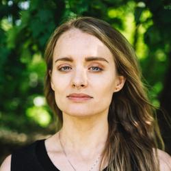 Kvinne med stripet brunt og blondt hår med blå øyne. Grønne trær i bakgrunnen.