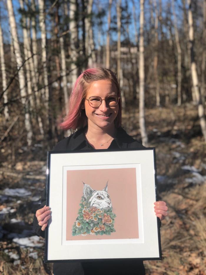 Bilde av kunsteren som står foran et skogholt og holder et av sine egne bilder; en tegning av en gaupe med blomster