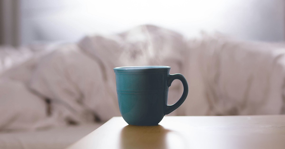 A cup of tea steaming on a bedside table with a bed in the background