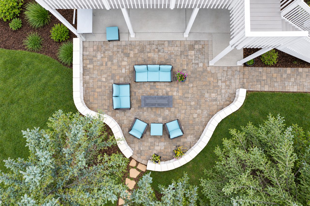Aerial view of a curved paver patio with outdoor seating beneath a white deck, bordered by lawn and evergreens.