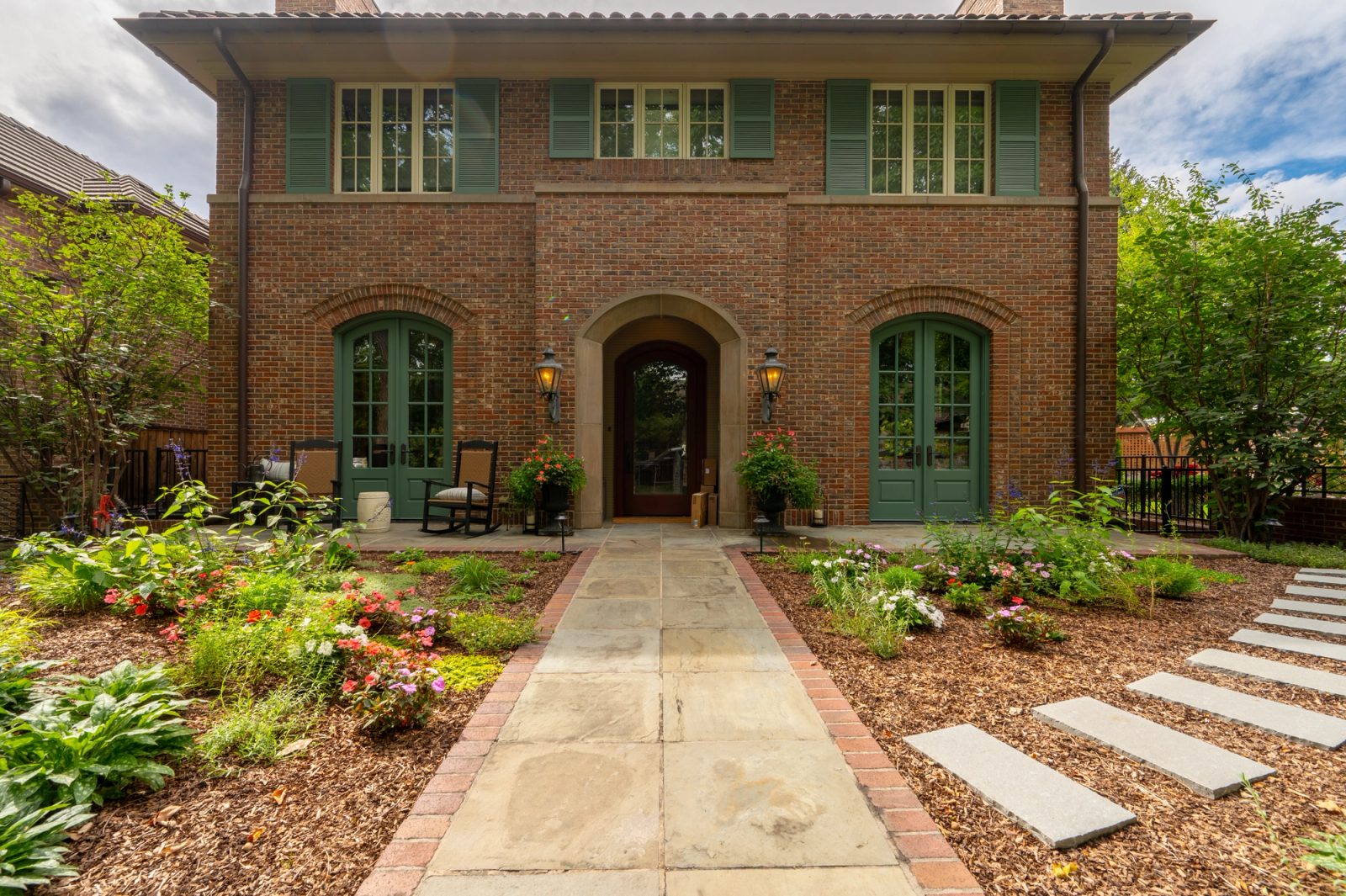 Brick-edged bluestone walk through lush perennial beds to the arched front door of a red-brick home with green shutters.