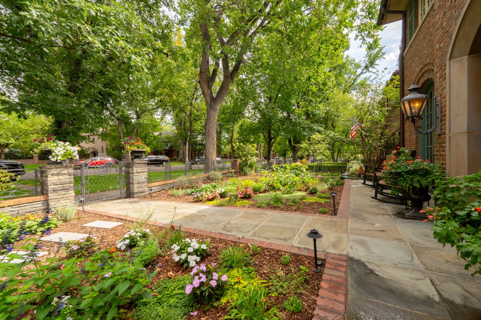 Flowering beds and flagstone walkway in a shaded front garden beside a brick house.