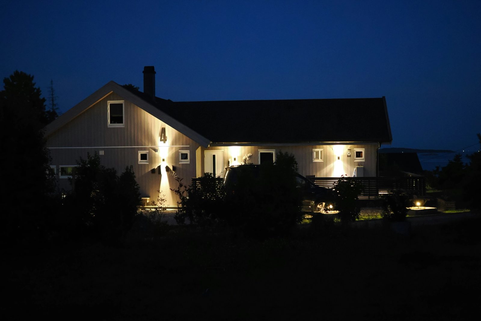 A two-story clapboard house at blue hour with warm lights washing its walls and garden.