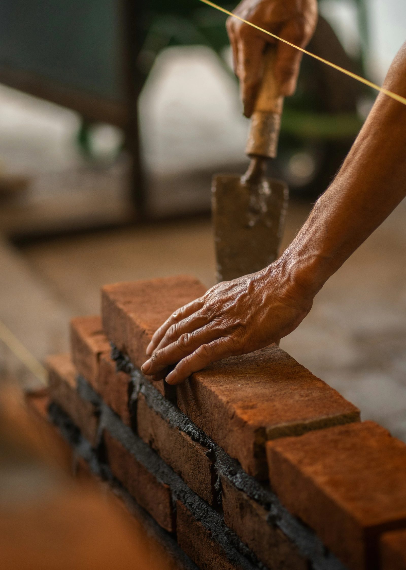 Weathered hands setting a red brick on a mortar bed along a taut string line, trowel in hand.