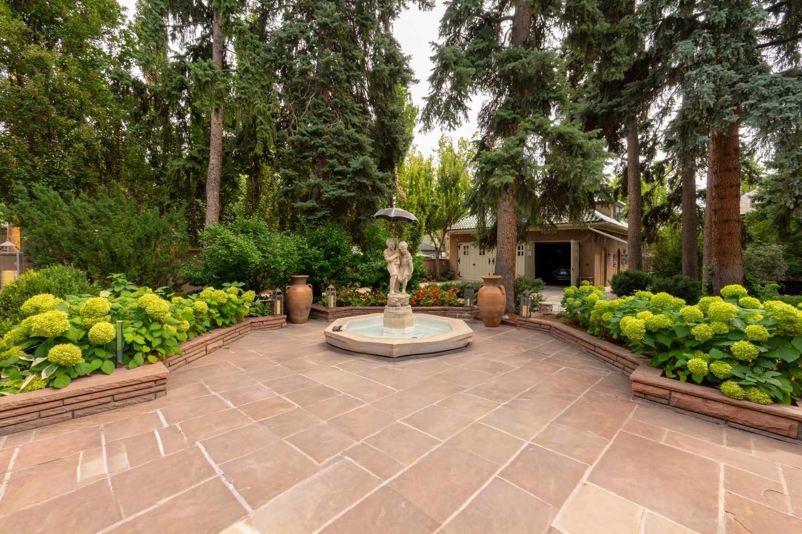 A statue fountain centers a red sandstone flagstone courtyard framed by hydrangea-filled stone planters and tall spruces.
