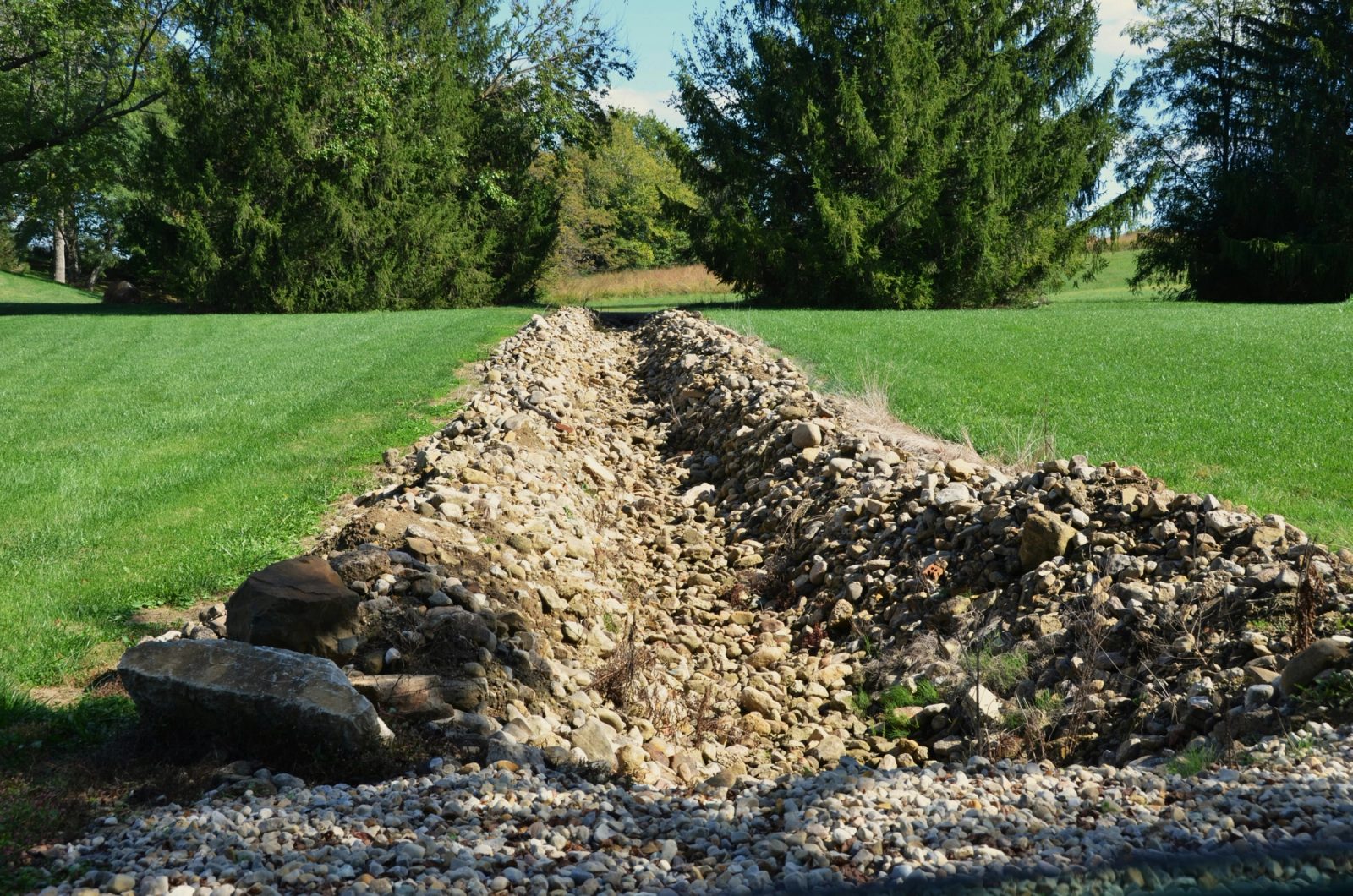 A freshly dug drainage trench lined with gravel and rock runs across a green residential lawn toward evergreen trees.