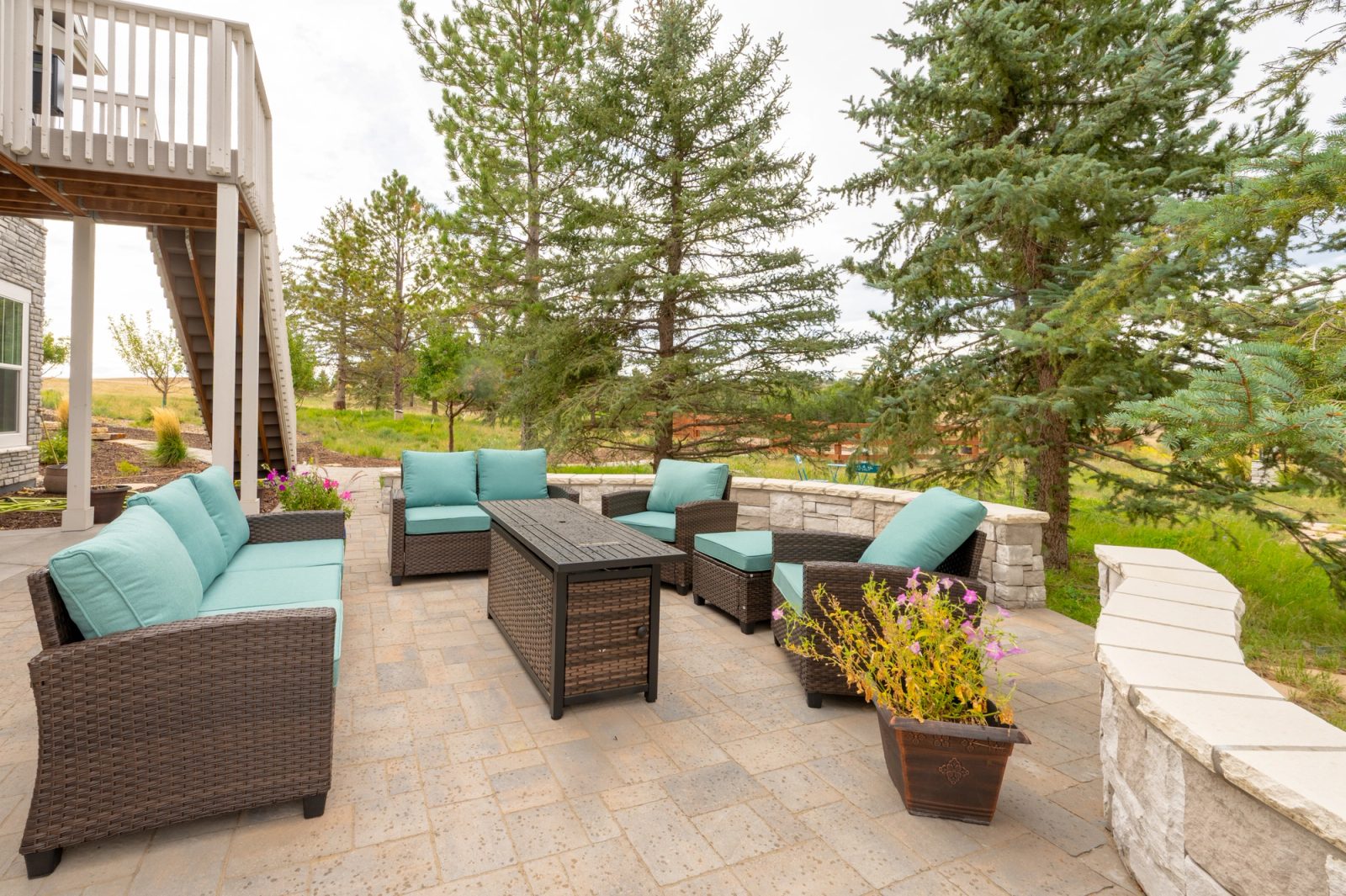 Paver patio with cushioned wicker furniture and a fire table beneath a deck, framed by tall pines.