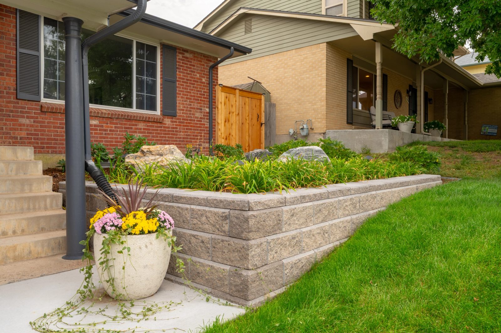 Tan split-face block retaining wall stepping up a lawn slope beside a front walk with a flowering planter.