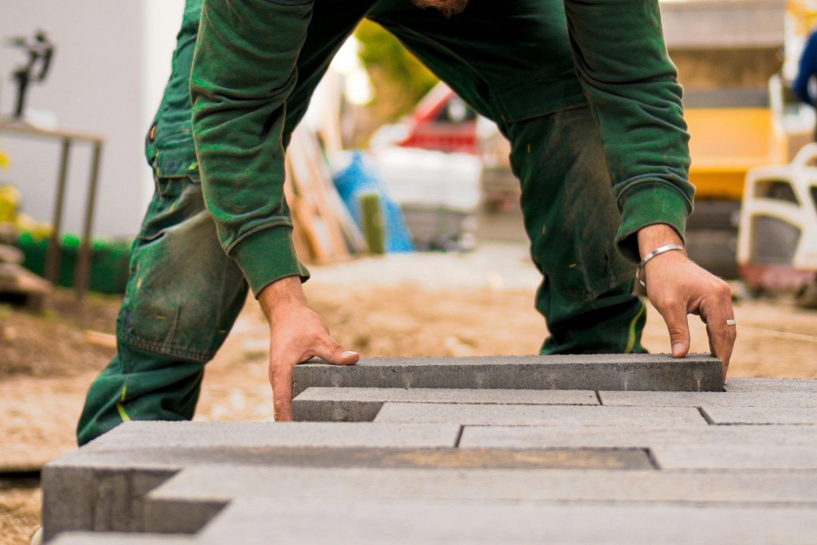 Hands of a worker in green workwear setting gray concrete pavers into a patio pattern.