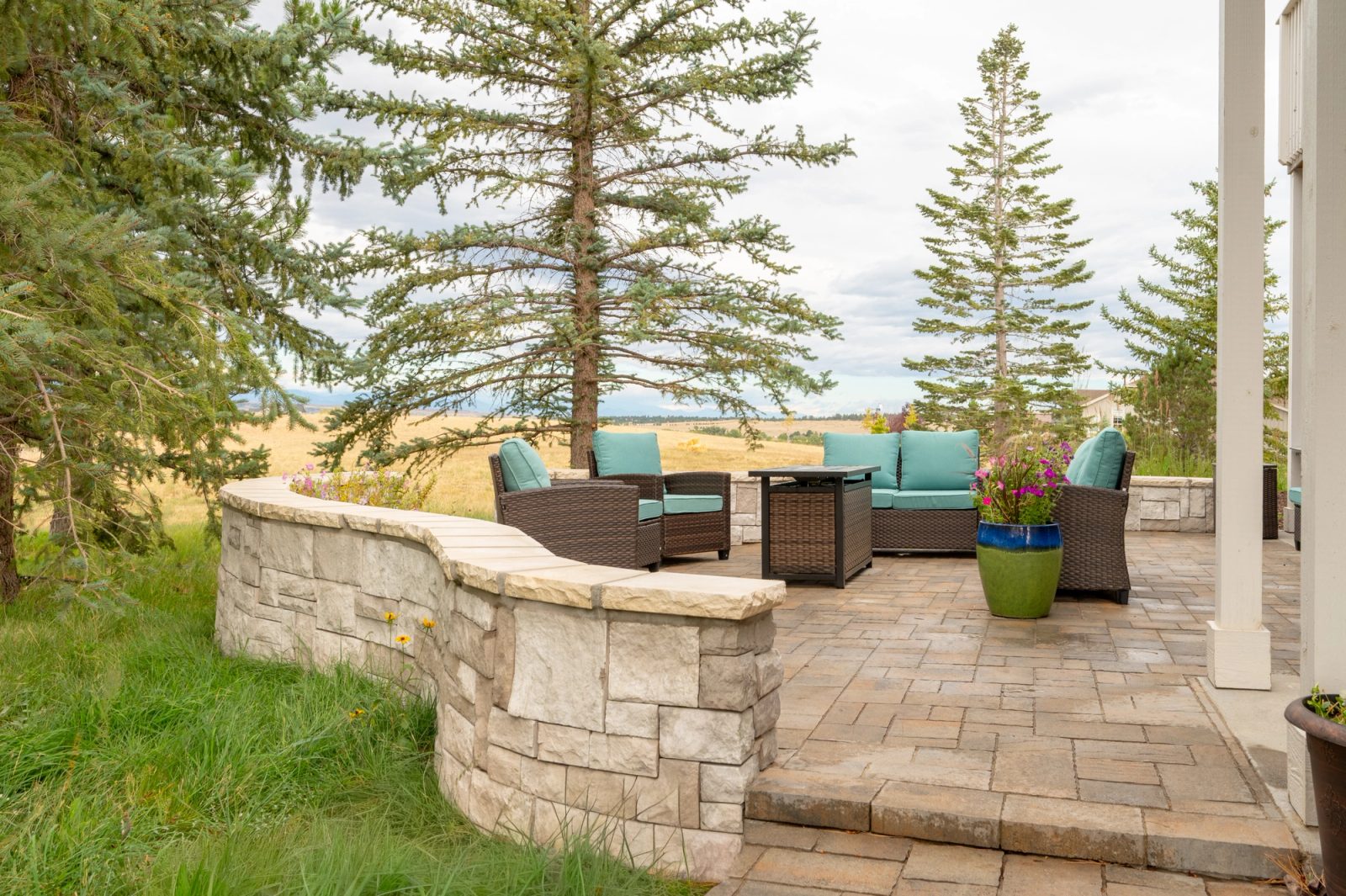 Curved stacked-stone seat wall enclosing a paver patio with seating, set among lawn and pines.