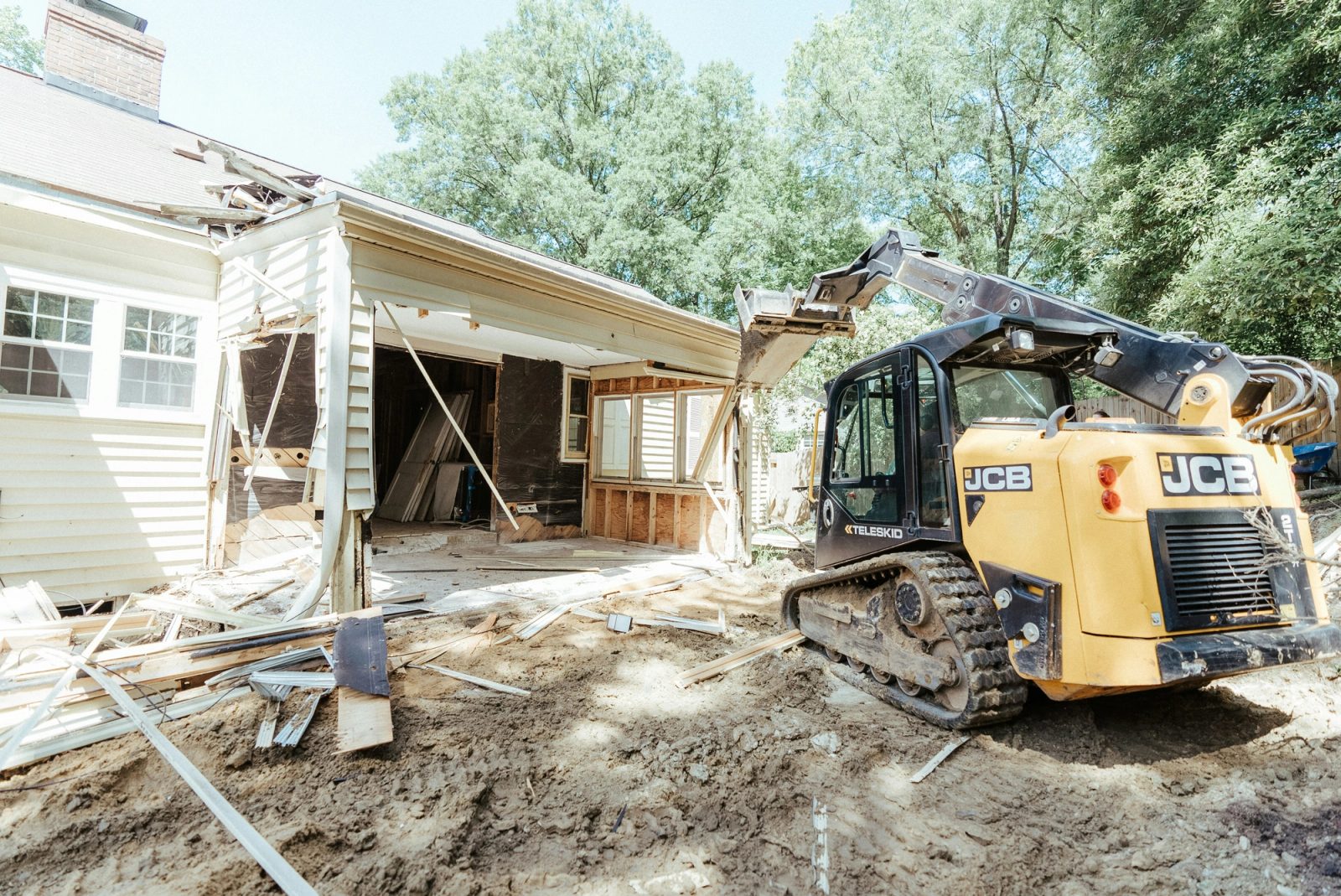 A compact track loader tearing out an old addition and clearing raw soil beside a white clapboard house.