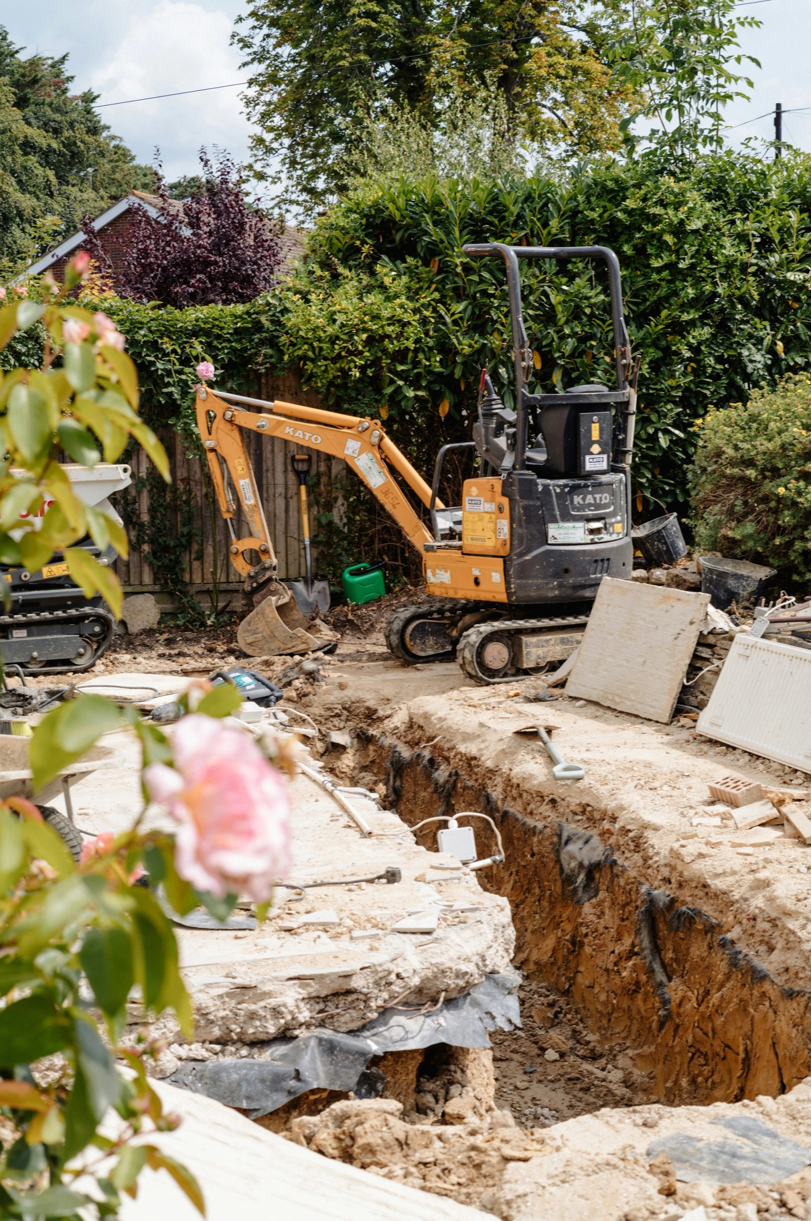 A small orange mini excavator beside a freshly dug trench in a residential backyard with a wooden fence and garden shrubs.