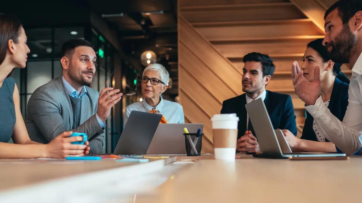 Six colleagues in a discussion around a conference table with laptops and coffee