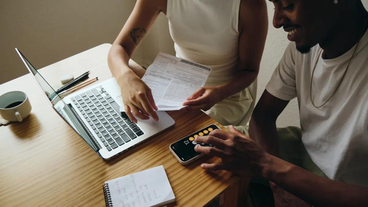 Two colleagues at a wooden desk comparing a printed form with figures on a laptop and a phone calculator
