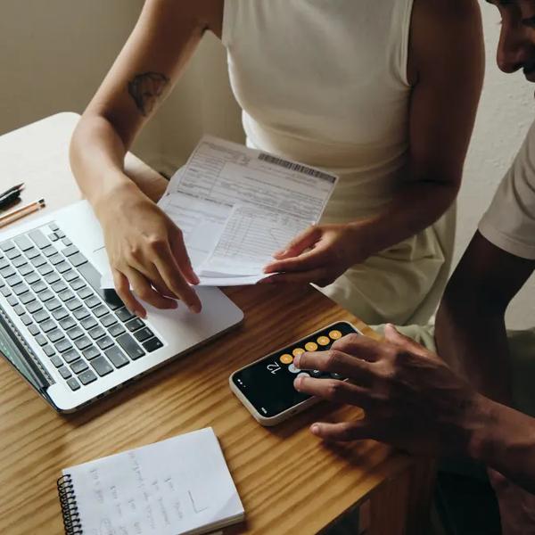 Two colleagues at a wooden desk comparing a printed form with figures on a laptop and a phone calculator