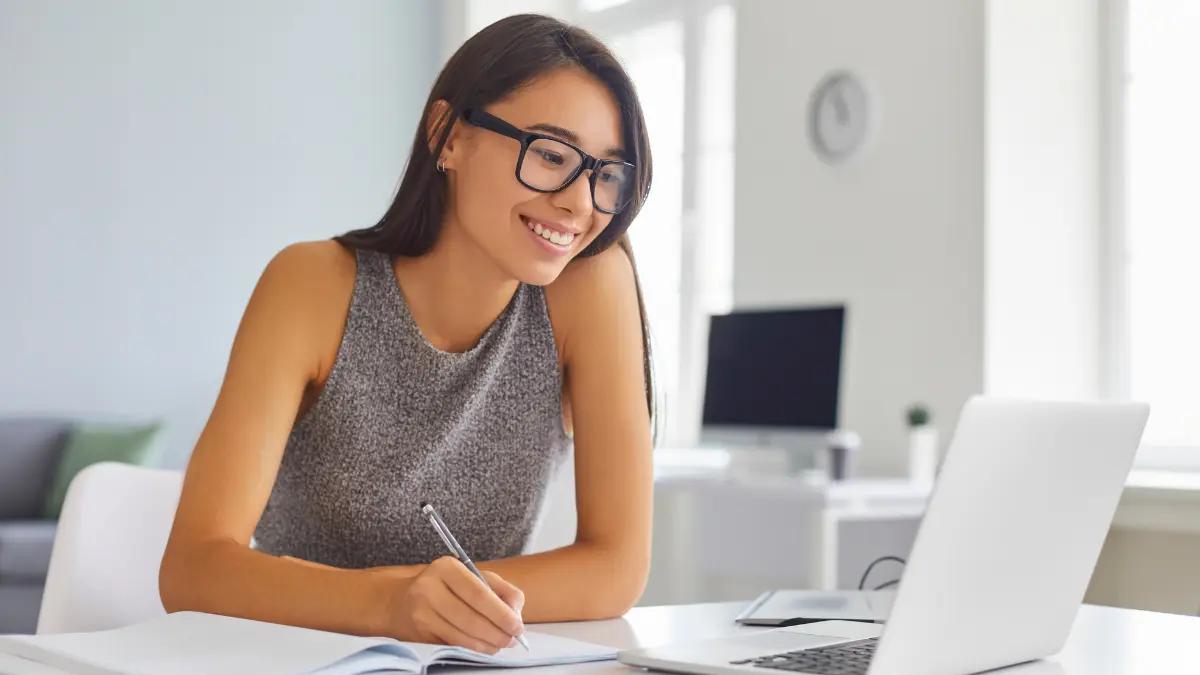 A woman in glasses writing notes in a notebook beside an open laptop