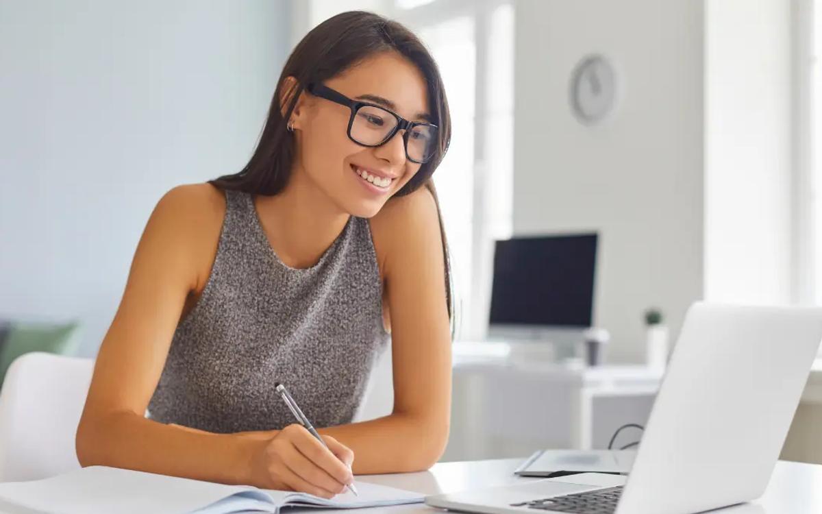A woman in glasses writing notes in a notebook beside an open laptop