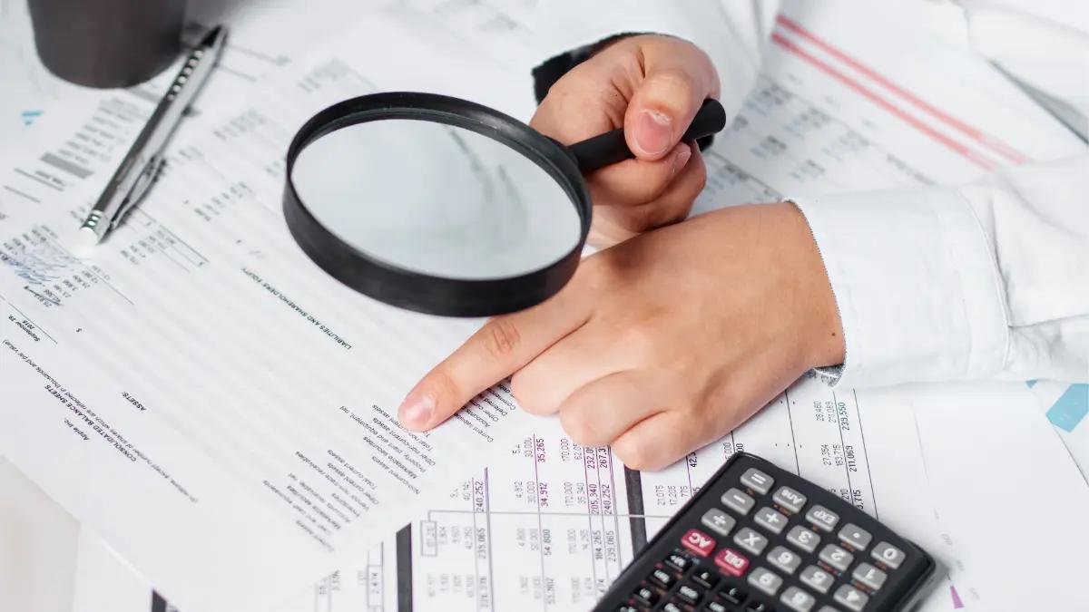 Hand holding a magnifying glass over printed spreadsheets next to a calculator