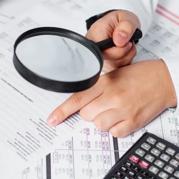 Hand holding a magnifying glass over printed spreadsheets next to a calculator