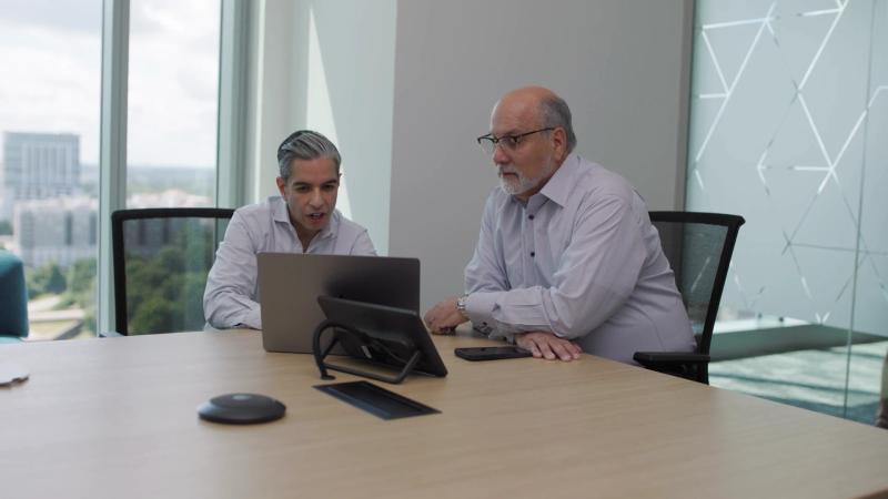 Two colleagues look at a laptop together at a conference table in an office overlooking the city