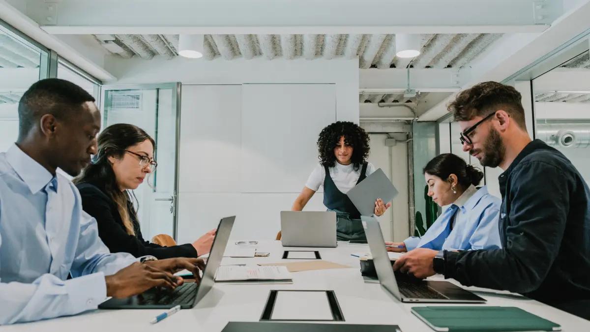 Five colleagues around a conference table with open laptops while one of them stands to present