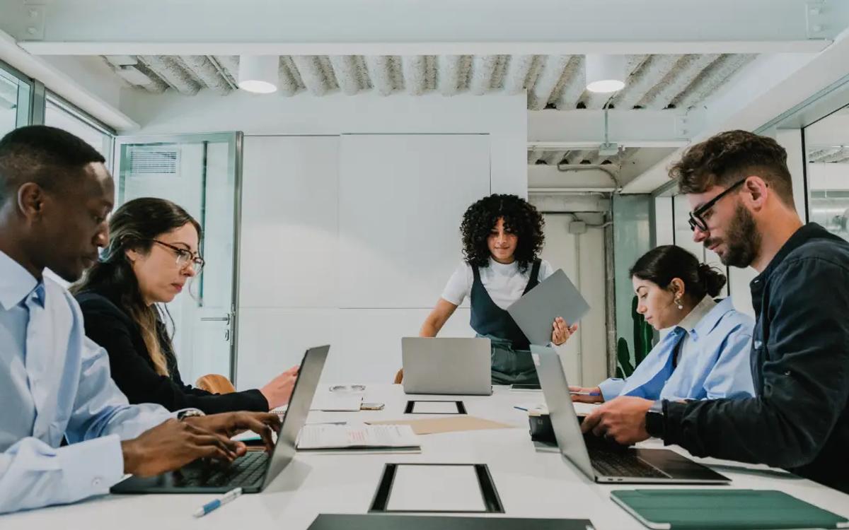 Five colleagues around a conference table with open laptops while one of them stands to present