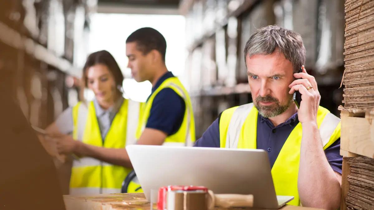 Three warehouse workers in high-visibility vests, one on the phone checking a laptop between pallets