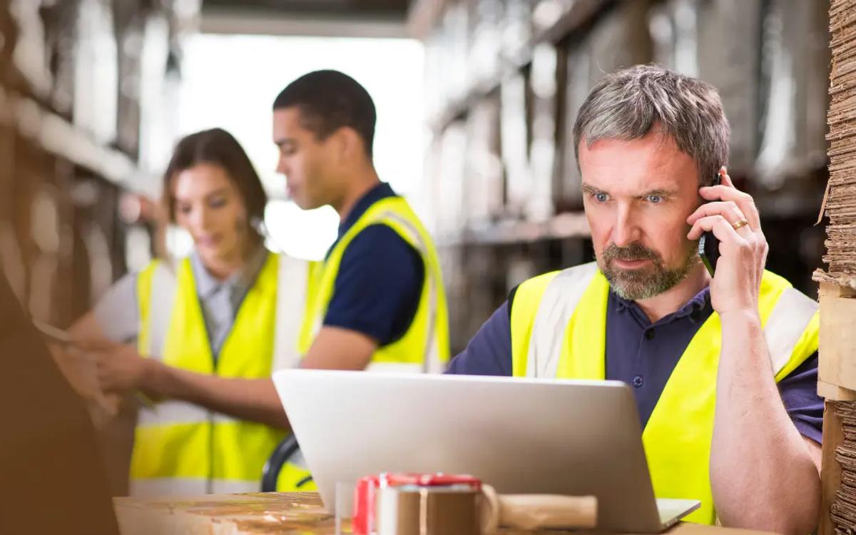 Three warehouse workers in high-visibility vests, one on the phone checking a laptop between pallets