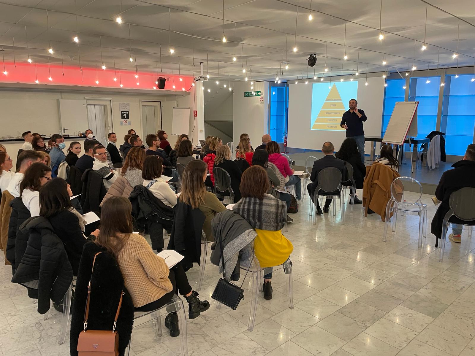 Uomo su un palco che parla di fronte ad un pubblico di persone sedute