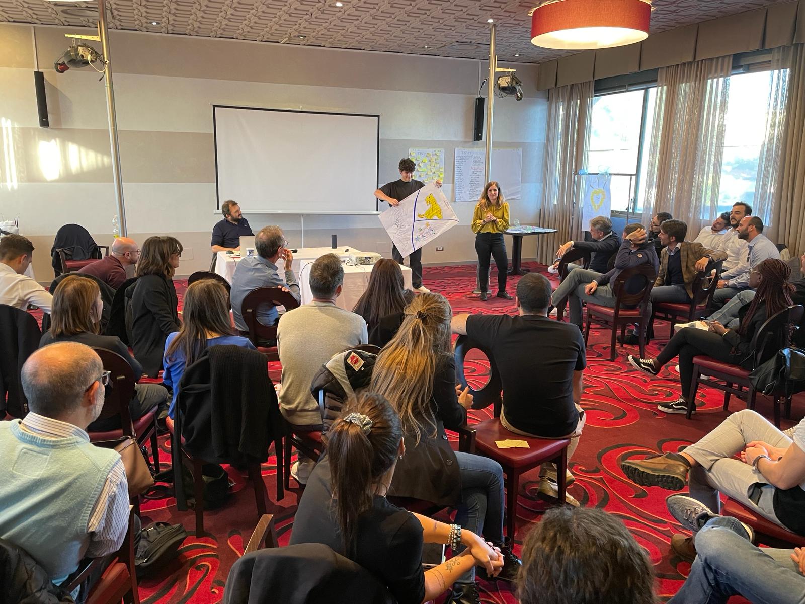 Due persone che presentano un progetto davanti ad un pubblico seduto