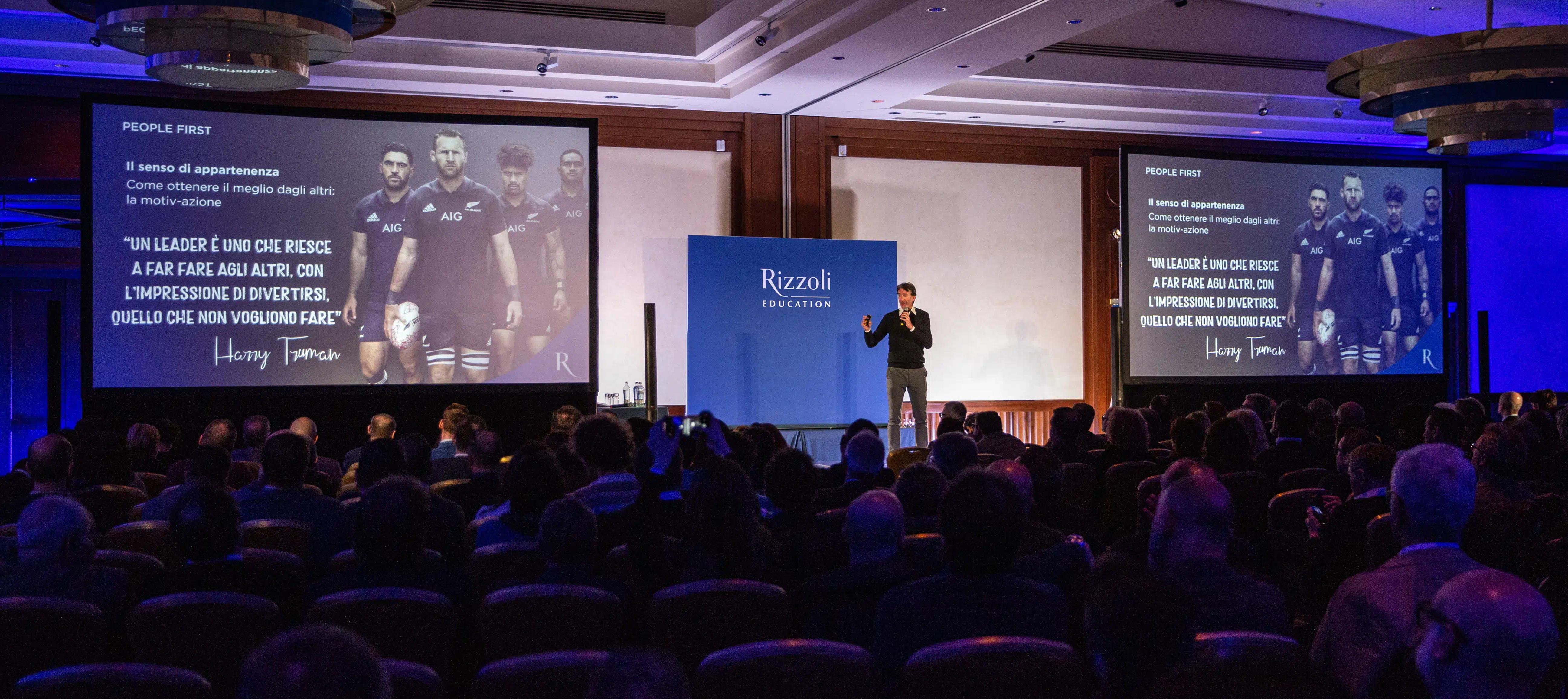 un gruppo di persone sedute davanti ad un palco con alcuni schermi illuminati