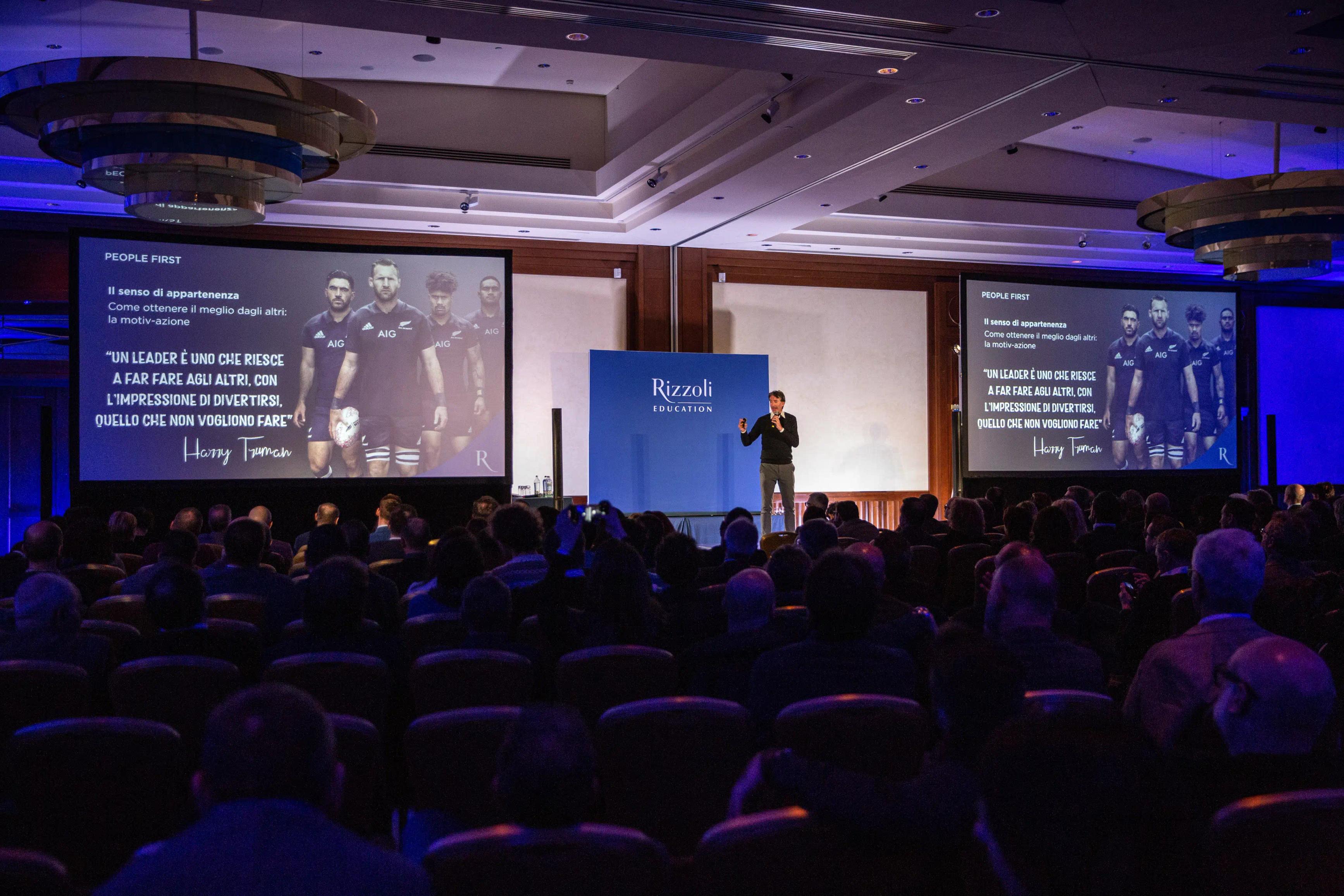 un gruppo di persone sedute davanti ad un palco con alcuni schermi illuminati