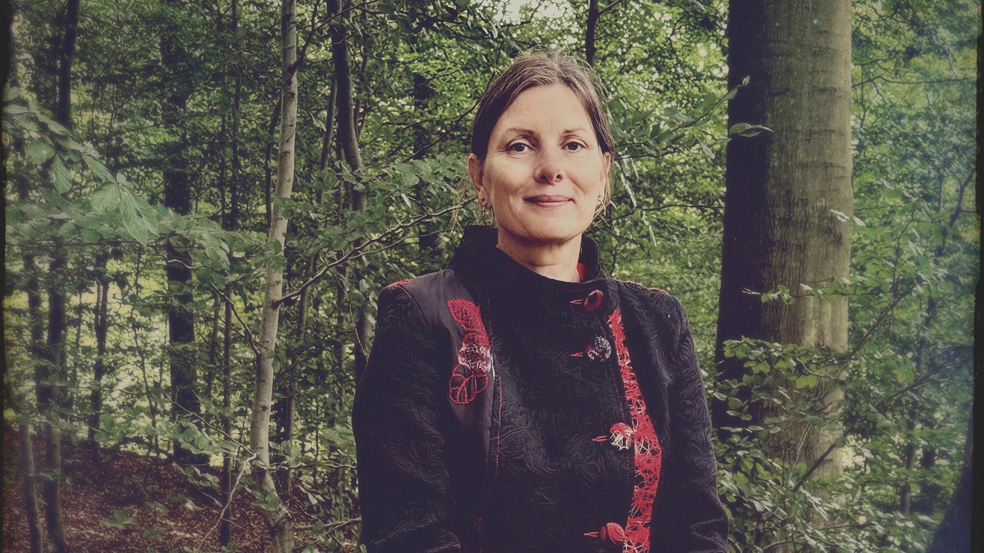 Zita Fejerskov Andersen står i et skovbryn i en mørk jakke med rødt broderi