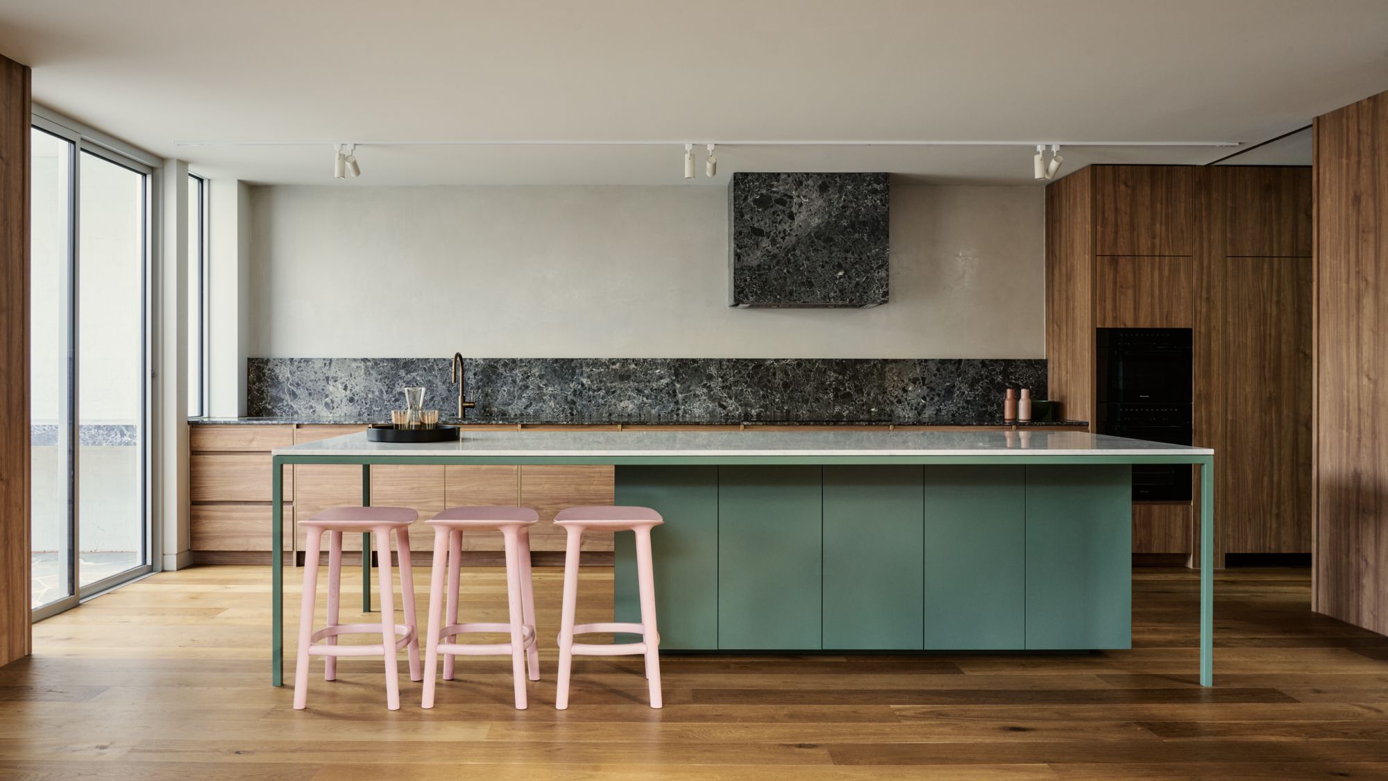 An elevation image of the full length of the kitchen with olive cabinets, pink bar stools and grey marble on the further benchtop and rangehood.| Peavey Residence II by FYC Architects and Snakes & Ladders Projects | Prahran | Veeral Patel